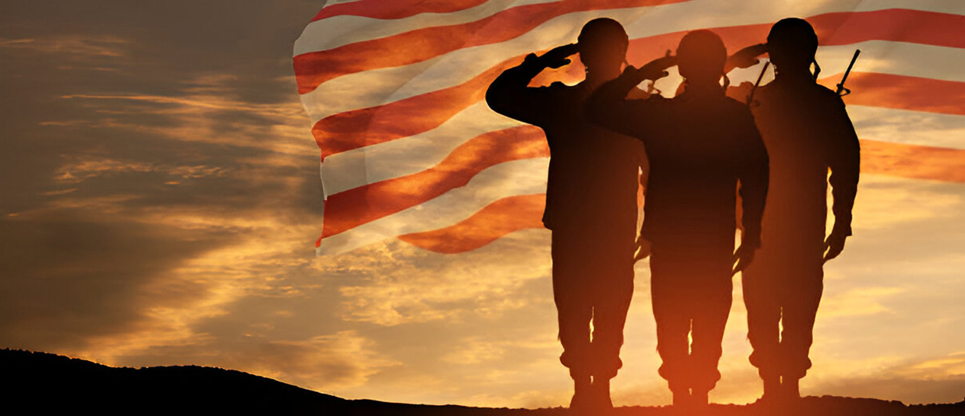 Three soldiers saluting at sunset with an American flag waving in the background