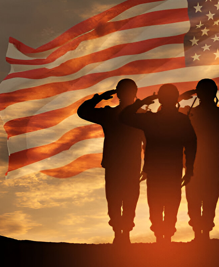 Three soldiers saluting at sunset with an American flag waving in the background