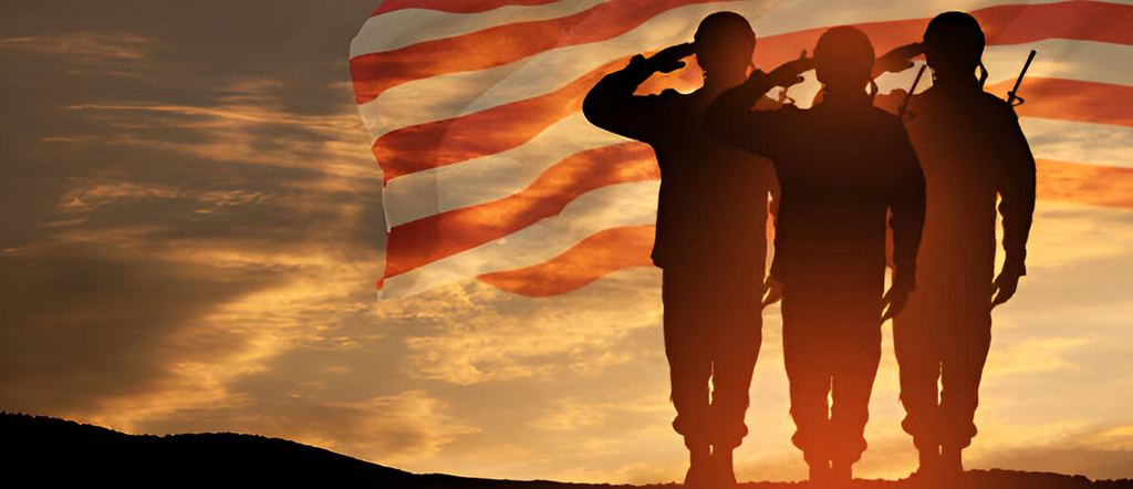 Three soldiers saluting at sunset with an American flag waving in the background